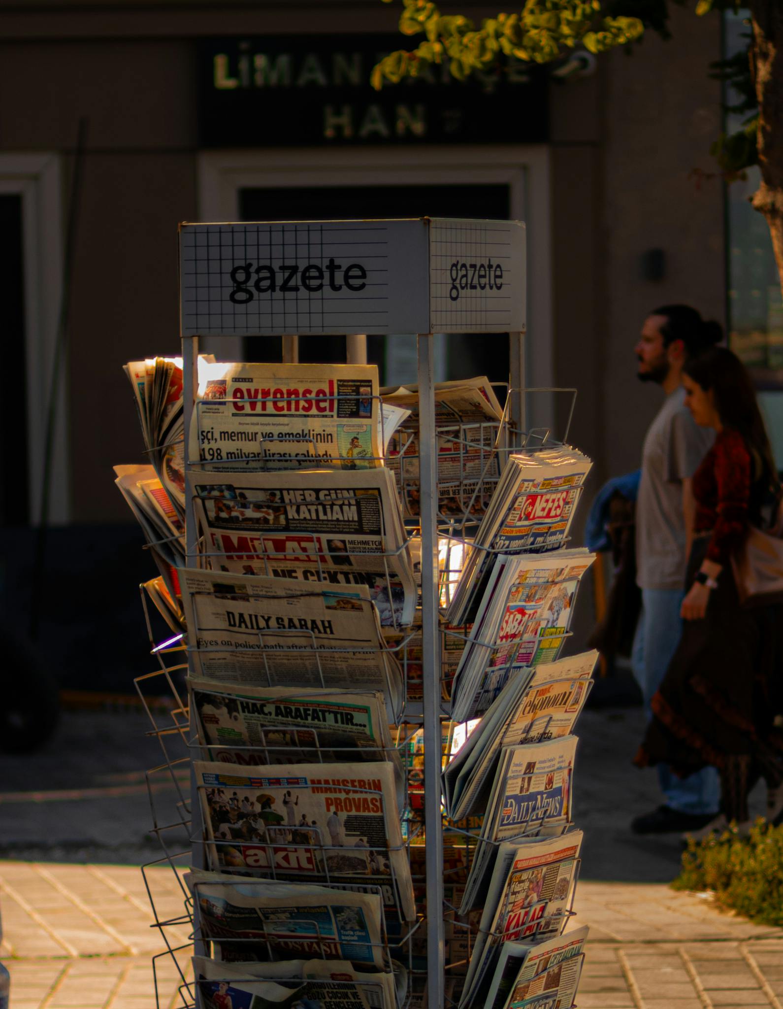 A newspaper stand with various publications on a sunny street corner.