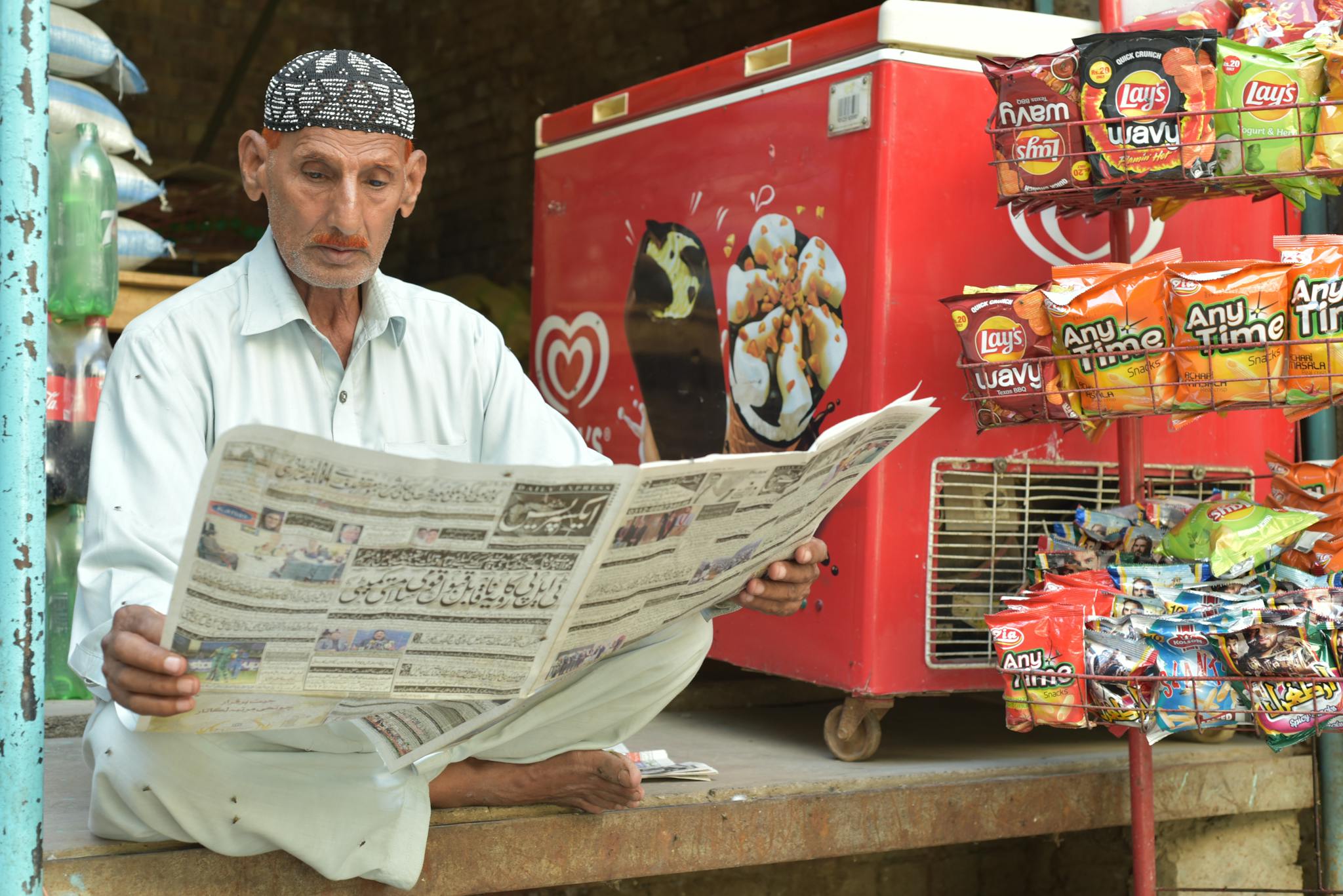 A senior man sits reading a newspaper at a small store in Multan, surrounded by snacks and a freezer.